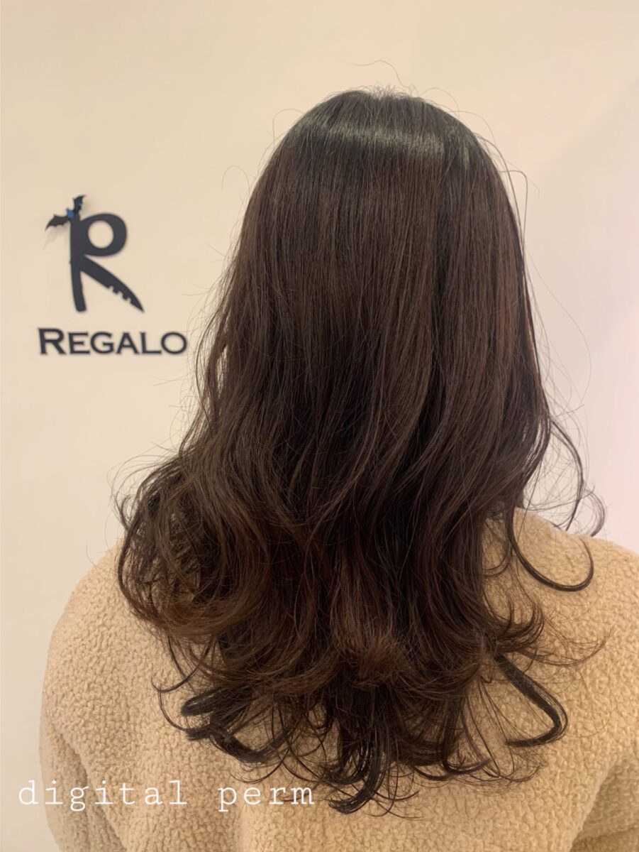 ベージュの壁の前で見せる秋色ブラウンの柔らかいケアデジタルパーマロングヘア後ろ姿