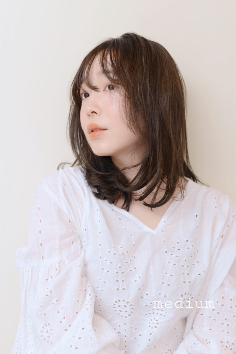 明るいベージュ背景でふんわり内巻きミディアムヘアと白レースブラウスの女性
